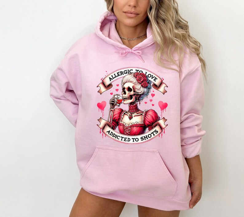 May include: Light pink hoodie with a front pocket. The graphic on the front features a skeleton in a red dress drinking from a glass, with the text "ALLERGIC TO LOVE" and "ADDICTED TO SHOTS" on banners.