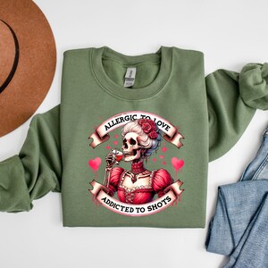 May include: An olive green sweatshirt featuring a graphic of a skeleton in a red dress, drinking from a glass. The graphic includes the text "Allergic to Love, Addicted to Shots" in a banner design, with pink hearts.