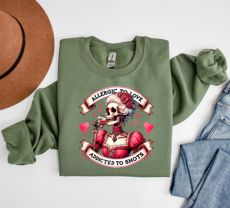 May include: An olive green sweatshirt featuring a graphic of a skeleton in a red dress, drinking from a glass. The graphic includes the text "Allergic to Love, Addicted to Shots" in a banner design, with pink hearts.