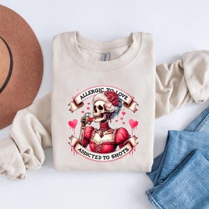 May include: Beige sweatshirt with a graphic of a skeleton in a red dress, holding a glass of red liquid. The graphic includes the text "ALLERGIC TO LOVE" and "ADDICTED TO SHOTS" with hearts. The sweatshirt is on a white surface with a brown hat and blue jeans.