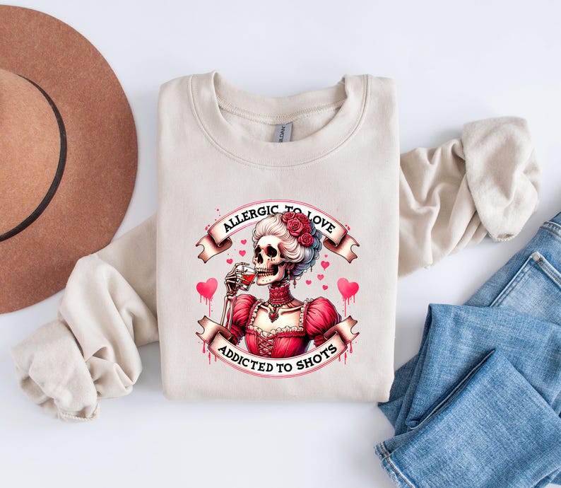 May include: Beige sweatshirt with a graphic of a skeleton in a red dress, holding a glass of red liquid. The graphic includes the text "ALLERGIC TO LOVE" and "ADDICTED TO SHOTS" with hearts. The sweatshirt is on a white surface with a brown hat and blue jeans.