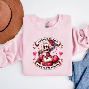 May include: A light pink sweatshirt featuring a skeleton illustration in a red dress, drinking from a glass. The text reads "Allergic to Love, Addicted to Shots." The design includes hearts and roses. A brown hat and blue jeans are in the background.
