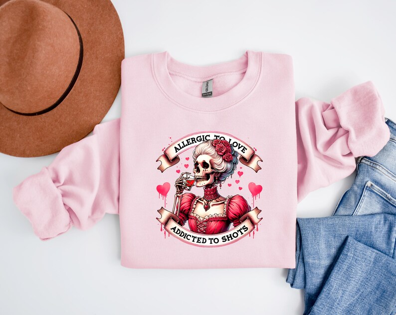 May include: A light pink sweatshirt featuring a skeleton illustration in a red dress, drinking from a glass. The text reads "Allergic to Love, Addicted to Shots." The design includes hearts and roses. A brown hat and blue jeans are in the background.