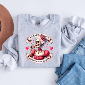 May include: A light gray sweatshirt featuring a graphic of a skeleton in a red dress, drinking from a glass. The text reads "Allergic to Love, Addicted to Shots" in a banner design. The design includes pink hearts.