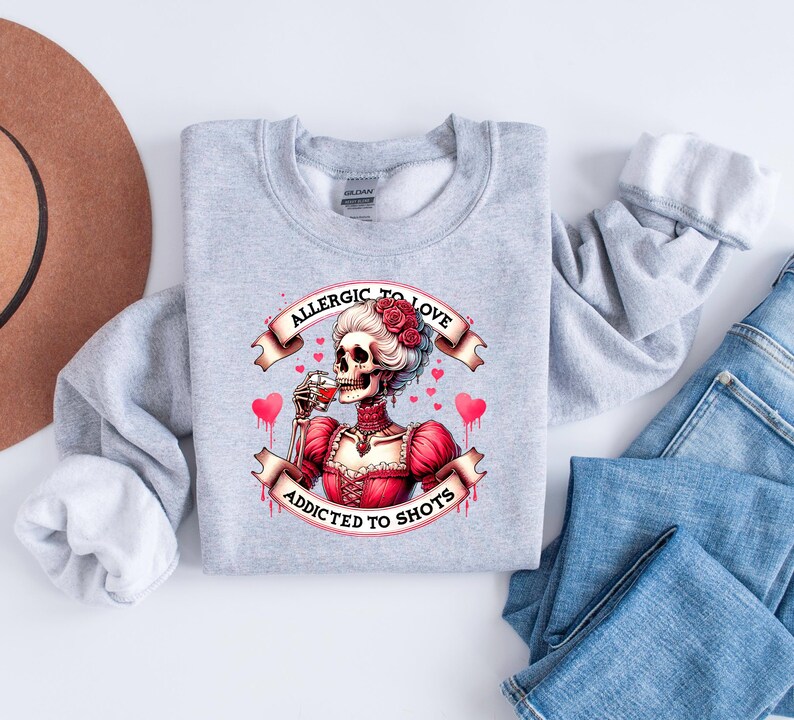 May include: A light gray sweatshirt featuring a graphic of a skeleton in a red dress, drinking from a glass. The text reads "Allergic to Love, Addicted to Shots" in a banner design. The design includes pink hearts.