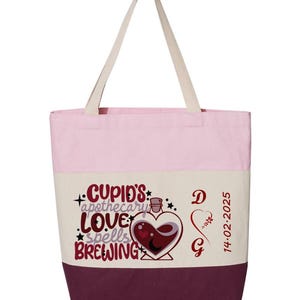 May include: A canvas tote bag with a pink top, cream middle, and burgundy bottom. The bag features the text "Cupid's Apothecary Love Spells Brewing" with a heart-shaped potion bottle graphic. The bag also has the initials "D G" and the date "14-02-2025".