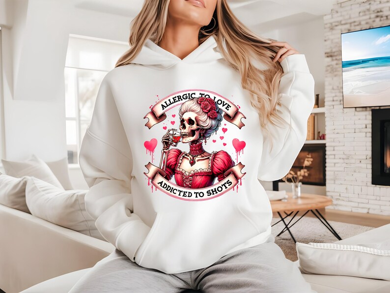May include: White hoodie featuring a graphic of a skeleton in a red dress, holding a glass of wine. The design includes the text "ALLERGIC TO LOVE" and "ADDICTED TO SHOTS" on banners, with pink hearts. The hoodie is a casual, comfortable garment.