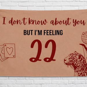 May include: A tan banner with the text "I don't know about you BUT I'M FEELING 22". The banner also features illustrations of playing cards, a leopard, and a globe. The text and illustrations are in a dark red color.
