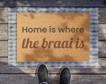 May include: A rectangular doormat with a tan center and a grey and white plaid border. The text "Home is where the braai is" is printed in dark brown letters across the center of the mat. The mat is positioned on a wooden deck surface.