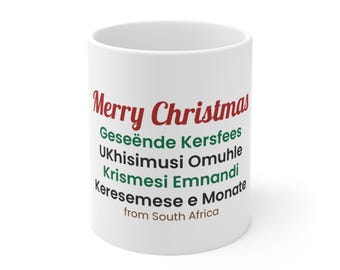 Op de afbeelding: Een witte keramische mok met de rode tekst Merry Christmas, gevolgd door Geseënde Kersfees, UKhisimusi Omuhle, Krismesi Emnandi en Keresemese e Monate in groen en zwart, met de bruine tekst from South Africa onderaan.
