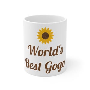 Könnte beinhalten: Ein weißer Keramikbecher mit einer gelben Sonnenblumen-Grafik über dem braunen Schriftzug "World's Best Gogo" in Schreibschrift. Der Becher hat einen glatten, zylindrischen Körper mit einer sauberen weißen Oberfläche und einem Standardhenkel.