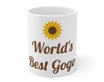 Może przedstawiać: Biały ceramiczny kubek z grafiką żółtego słonecznika umieszczoną nad brązowym napisem "World's Best Gogo" wykonanym kursywą. Kubek ma gładki, cylindryczny korpus z czystym białym wykończeniem oraz standardowy uchwyt z boku.