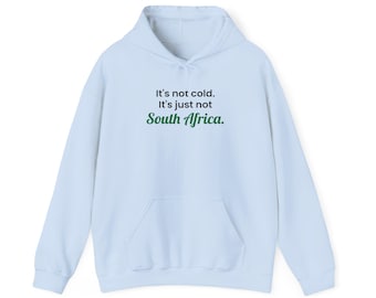 May include: A light blue hooded sweatshirt featuring a front kangaroo pocket and long sleeves. The chest displays the black text "It's not cold. It's just not" followed by the word "South Africa." in a green cursive script font.
