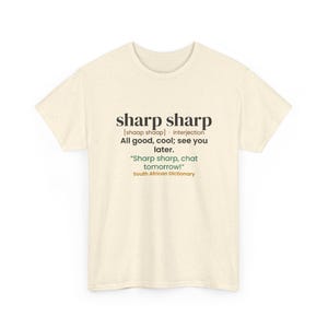 May include: A cream-colored short-sleeve t-shirt with a speckled texture. The front features black text defining the phrase sharp sharp as an interjection meaning all good or cool, followed by the example sentence Sharp sharp, chat tomorrow! and the text South African Dictionary in orange.
