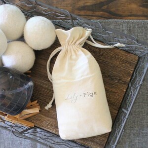 Puede incluir: Una bolsa crema con cord&oacute;n y el texto "Lily + Figs" se encuentra sobre una bandeja de madera, junto a varias bolas blancas de lana para secadora. La bandeja est&aacute; dentro de una cesta de alambre met&aacute;lico, con un fondo de tela gris.