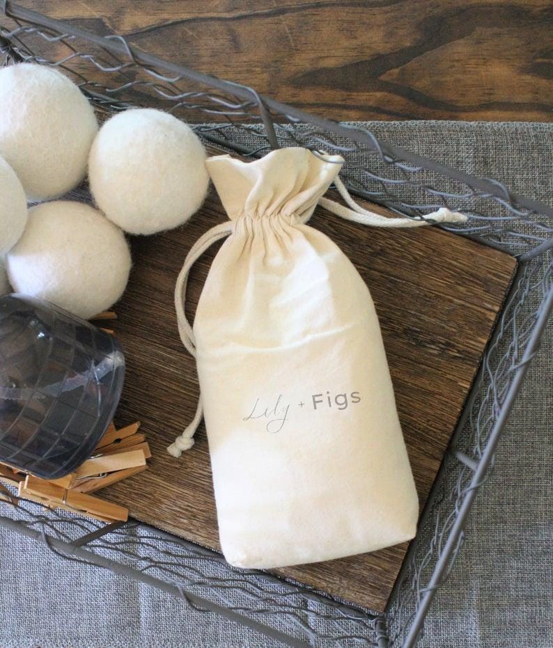 Puede incluir: Una bolsa crema con cord&oacute;n y el texto "Lily + Figs" se encuentra sobre una bandeja de madera, junto a varias bolas blancas de lana para secadora. La bandeja est&aacute; dentro de una cesta de alambre met&aacute;lico, con un fondo de tela gris.