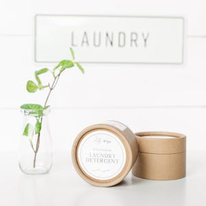 Può includere: Un vaso di vetro trasparente con una pianta verde e due contenitori di cartone marrone di detersivo per bucato. L'etichetta sul detersivo recita "LAUNDRY DETERGENT" e "Lily + Figs". Un cartello bianco con la parola "LAUNDRY" è sullo sfondo.