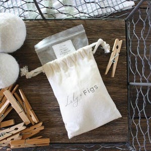 May include: A collection of laundry essentials, including white wool dryer balls, wooden clothespins, and a drawstring bag with the text "Lily + Figs." The items are arranged on a wooden surface, with a wire basket in the background.