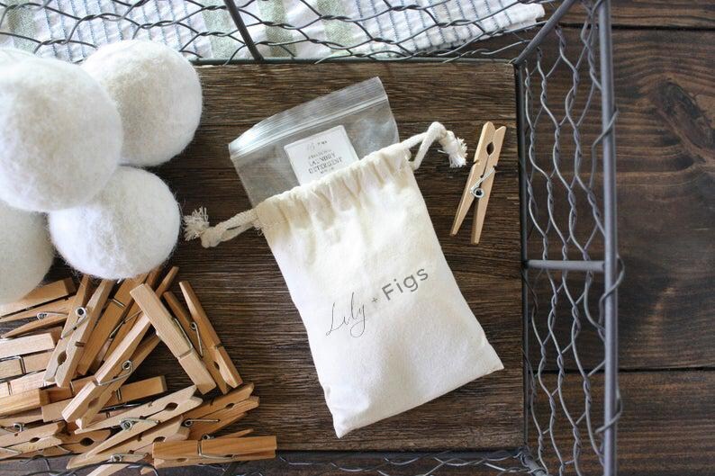 May include: A collection of laundry essentials, including white wool dryer balls, wooden clothespins, and a drawstring bag with the text "Lily + Figs." The items are arranged on a wooden surface, with a wire basket in the background.