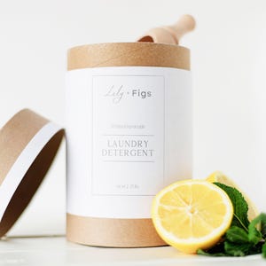 Op de afbeelding: Een cilindrische container met wasmiddel en een deksel, met het opschrift "Lily + Figs" en "Laundry Detergent". Een houten schepje ligt erin. Een halve citroen en muntblaadjes liggen ervoor.