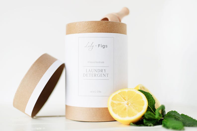 Op de afbeelding: Een cilindrische container met wasmiddel en een deksel, met het opschrift "Lily + Figs" en "Laundry Detergent". Een houten schepje ligt erin. Een halve citroen en muntblaadjes liggen ervoor.