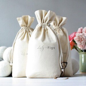 May include: Off-white drawstring bags with the text "Lily + Figs" printed on them. The bags are gathered at the top and have a natural, textured appearance. Several white wool dryer balls are in front of the bags.