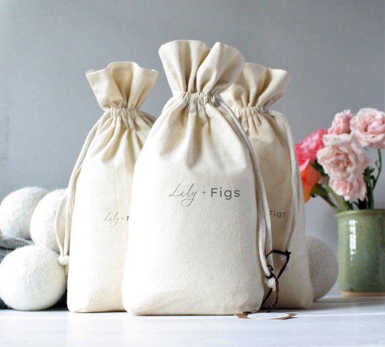 May include: Off-white drawstring bags with the text "Lily + Figs" printed on them. The bags are gathered at the top and have a natural, textured appearance. Several white wool dryer balls are in front of the bags.