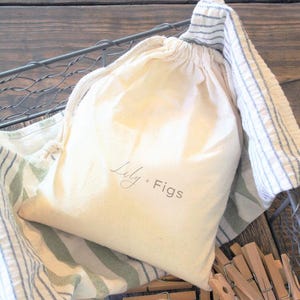 Puede incluir: Una bolsa crema con cord&oacute;n, impresa con "Lily + Figs", descansa en una cesta de alambre forrada con tela a rayas. Se ven pinzas de madera.
