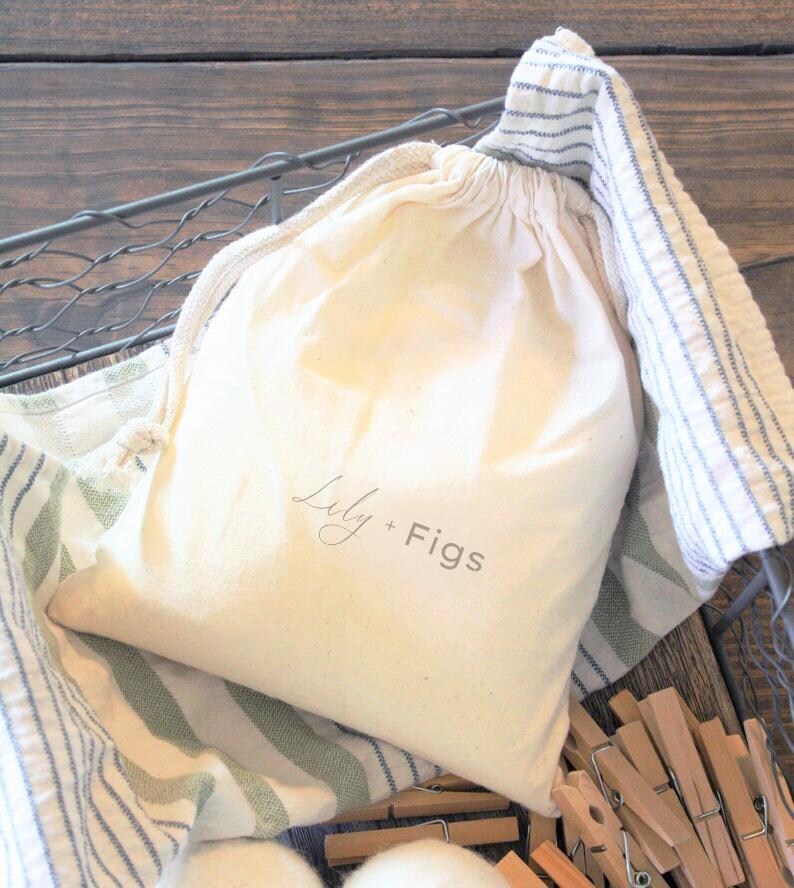 Puede incluir: Una bolsa crema con cord&oacute;n, impresa con "Lily + Figs", descansa en una cesta de alambre forrada con tela a rayas. Se ven pinzas de madera.
