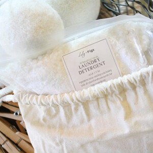 Puede incluir: Una bolsa de lona blanca llena de detergente para ropa, una bolsa de pl&aacute;stico transparente de detergente y tres bolas de secadora de lana blanca. La bolsa tiene un cierre de cord&oacute;n y una etiqueta que dice "Lily + Figs Laundry Detergent". Pinzas de ropa de madera en el fondo.