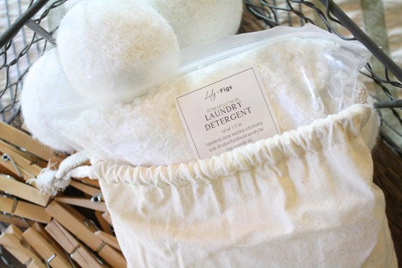 Puede incluir: Una bolsa de lona blanca llena de detergente para ropa, una bolsa de pl&aacute;stico transparente de detergente y tres bolas de secadora de lana blanca. La bolsa tiene un cierre de cord&oacute;n y una etiqueta que dice "Lily + Figs Laundry Detergent". Pinzas de ropa de madera en el fondo.