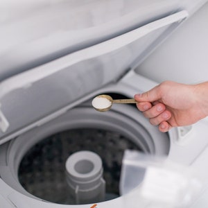 May include: A person adding laundry detergent to a white washing machine. A gold-colored measuring spoon holds a scoop of white powder. The washing machine lid is open, revealing the interior.
