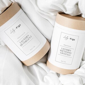 Op de afbeelding: Twee cilindrische containers met wasproducten liggen op witte stof. De containers zijn gemaakt van bruin karton met witte etiketten. Op &eacute;&eacute;n staat "Laundry Detergent" en op de andere "Laundry Softening Crystals". De merknaam "Lily. Figs" is ook zichtbaar.