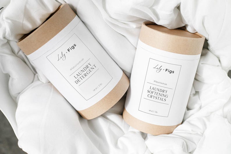 Op de afbeelding: Twee cilindrische containers met wasproducten liggen op witte stof. De containers zijn gemaakt van bruin karton met witte etiketten. Op &eacute;&eacute;n staat "Laundry Detergent" en op de andere "Laundry Softening Crystals". De merknaam "Lily. Figs" is ook zichtbaar.