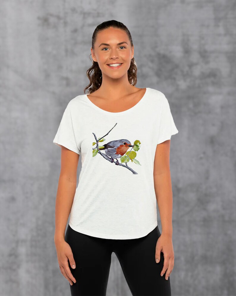 Puede incluir: Camiseta blanca con una ilustraci&oacute;n de acuarela de un petirrojo posado en una rama con hojas verdes.