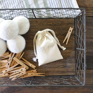 May include: A wire basket holds white felt balls, wooden clothespins, and a small drawstring bag with the text "Lily Figs." The items rest on a wooden surface, with a striped cloth in the background.