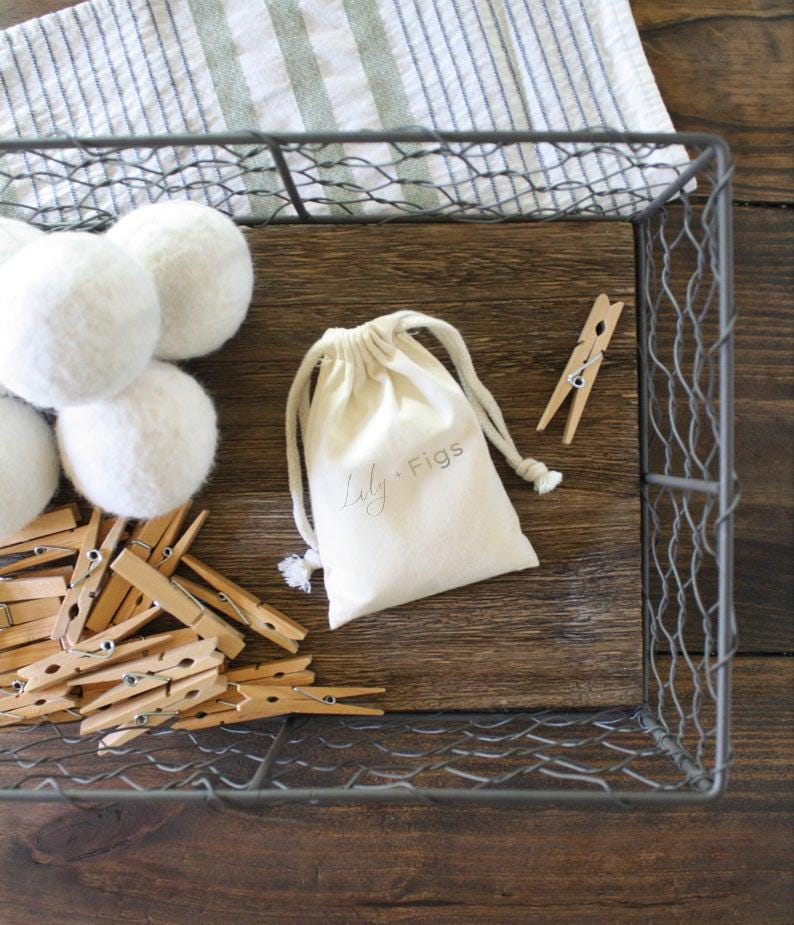 May include: A wire basket holds white felt balls, wooden clothespins, and a small drawstring bag with the text "Lily Figs." The items rest on a wooden surface, with a striped cloth in the background.