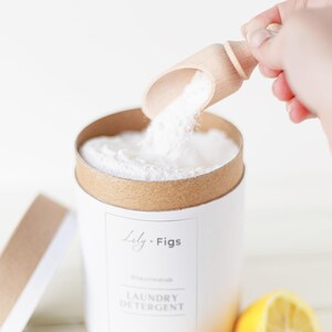 Op de afbeelding: Een witte cilindrische container met wasmiddel, met een bruine rand en deksel. Een houten schep giet wit poeder in de container. Op het etiket staat "Lily + Figs, Laundry Detergent". Twee gele citroenhelften staan op de voorgrond.