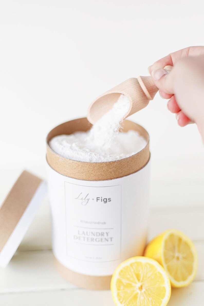 Op de afbeelding: Een witte cilindrische container met wasmiddel, met een bruine rand en deksel. Een houten schep giet wit poeder in de container. Op het etiket staat "Lily + Figs, Laundry Detergent". Twee gele citroenhelften staan op de voorgrond.