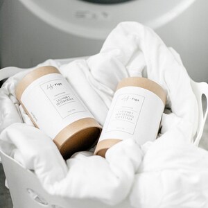 Op de afbeelding: Twee cilindrische containers met wasproducten, met het opschrift "Laundry Detergent" en "Laundry Softening Crystals", staan in een witte mand gevuld met wit linnen. De containers hebben een bruine basis en een wit etiket met de merknaam "Lily + Figs."