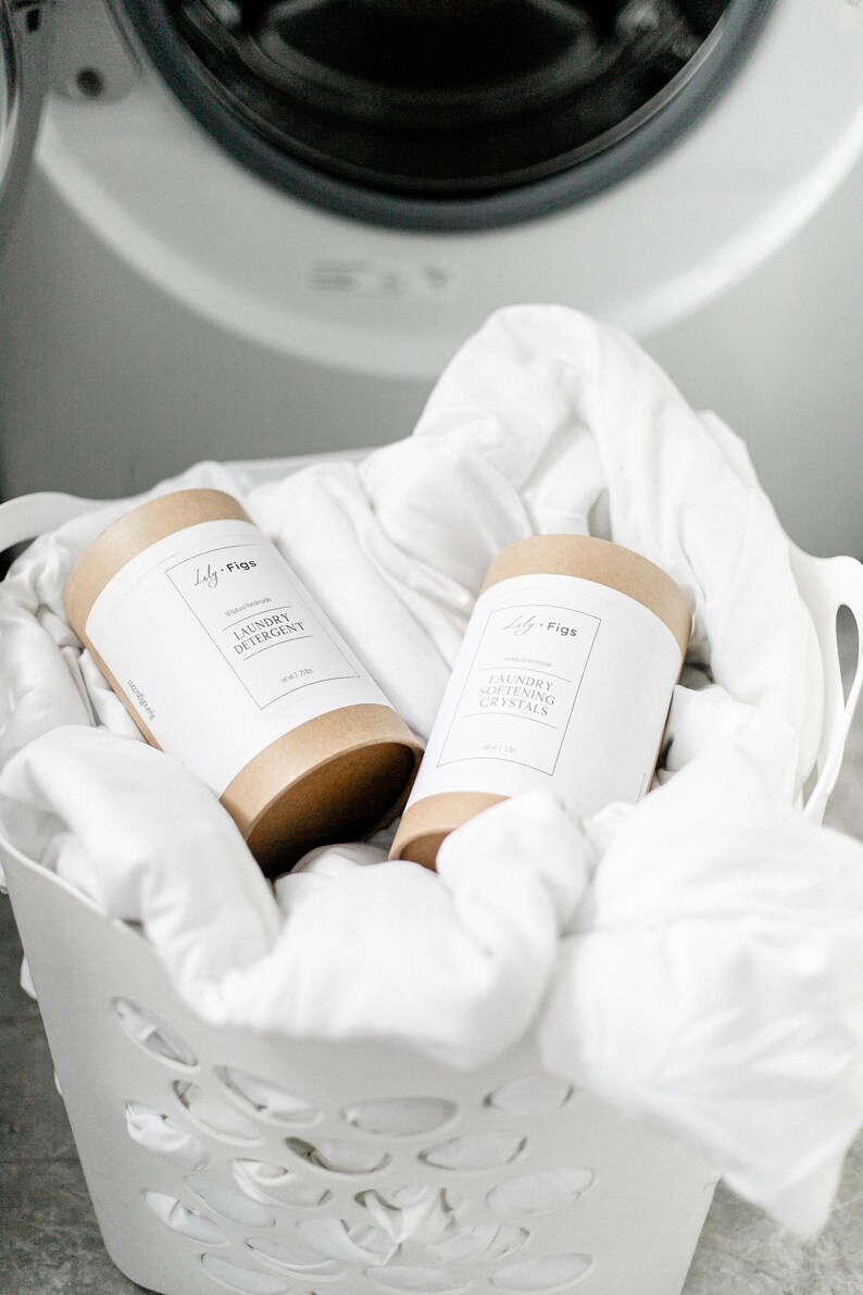 Op de afbeelding: Twee cilindrische containers met wasproducten, met het opschrift "Laundry Detergent" en "Laundry Softening Crystals", staan in een witte mand gevuld met wit linnen. De containers hebben een bruine basis en een wit etiket met de merknaam "Lily + Figs."