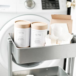 Op de afbeelding: Een metalen rolwagen bevat wasproducten. Twee witte canisters met bruine deksels zijn gelabeld met "Waspoeder" en "Wasverzachtende kristallen". Andere items zijn een stuk zeep, wasverzachterdoekjes en een wasbal.