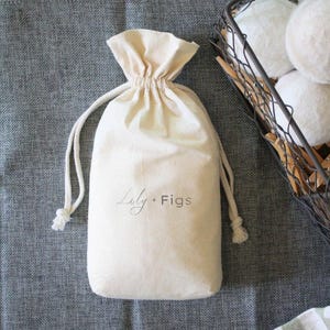 May include: A cream-colored drawstring bag with the text "Lily + Figs" in a script font. The bag is on a gray surface, with a wire basket filled with white spheres in the background.