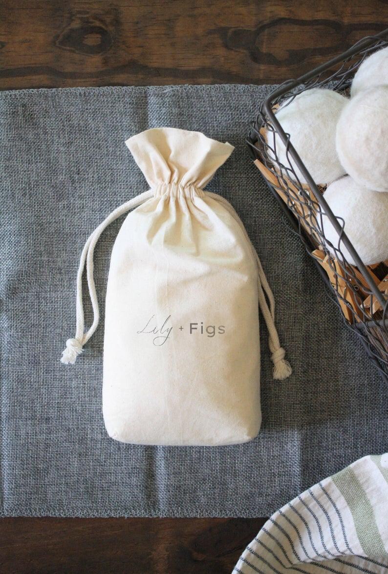 May include: A cream-colored drawstring bag with the text "Lily + Figs" in a script font. The bag is on a gray surface, with a wire basket filled with white spheres in the background.