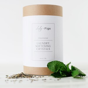 Op de afbeelding: Een cilindrische container met wasverzachtende kristallen van Lily + Figs, met een wit etiket en bruine boven- en onderkant. Op het etiket staat "LAUNDRY SOFTENING CRYSTALS" en "net 1.13 kg." Een takje munt en gedroogde kruiden liggen ervoor.