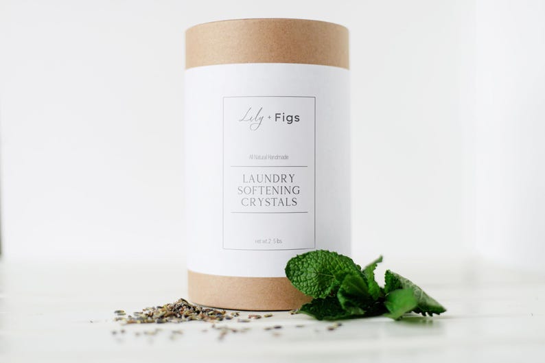 Op de afbeelding: Een cilindrische container met wasverzachtende kristallen van Lily + Figs, met een wit etiket en bruine boven- en onderkant. Op het etiket staat "LAUNDRY SOFTENING CRYSTALS" en "net 1.13 kg." Een takje munt en gedroogde kruiden liggen ervoor.