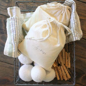May include: A wire basket filled with laundry essentials. It contains several white felted wool dryer balls, drawstring bags with "Lily. Figs" printed on them, wooden clothespins, and striped towels. The basket sits on a wooden surface.