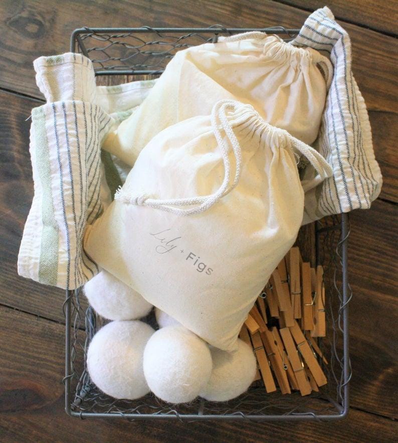 May include: A wire basket filled with laundry essentials. It contains several white felted wool dryer balls, drawstring bags with "Lily. Figs" printed on them, wooden clothespins, and striped towels. The basket sits on a wooden surface.