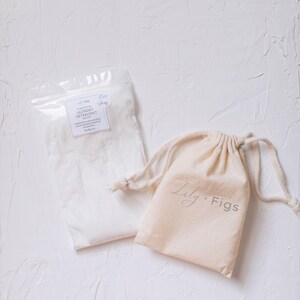 Puede incluir: Una bolsa de pl&aacute;stico transparente llena de detergente blanco para la ropa y una peque&ntilde;a bolsa beige con cord&oacute;n. La bolsa est&aacute; etiquetada como "Laundry Detergent" y la bolsa con cord&oacute;n tiene el texto "Lily + Figs" impreso.
