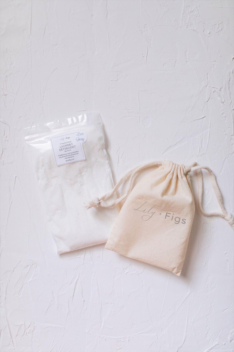 Puede incluir: Una bolsa de pl&aacute;stico transparente llena de detergente blanco para la ropa y una peque&ntilde;a bolsa beige con cord&oacute;n. La bolsa est&aacute; etiquetada como "Laundry Detergent" y la bolsa con cord&oacute;n tiene el texto "Lily + Figs" impreso.